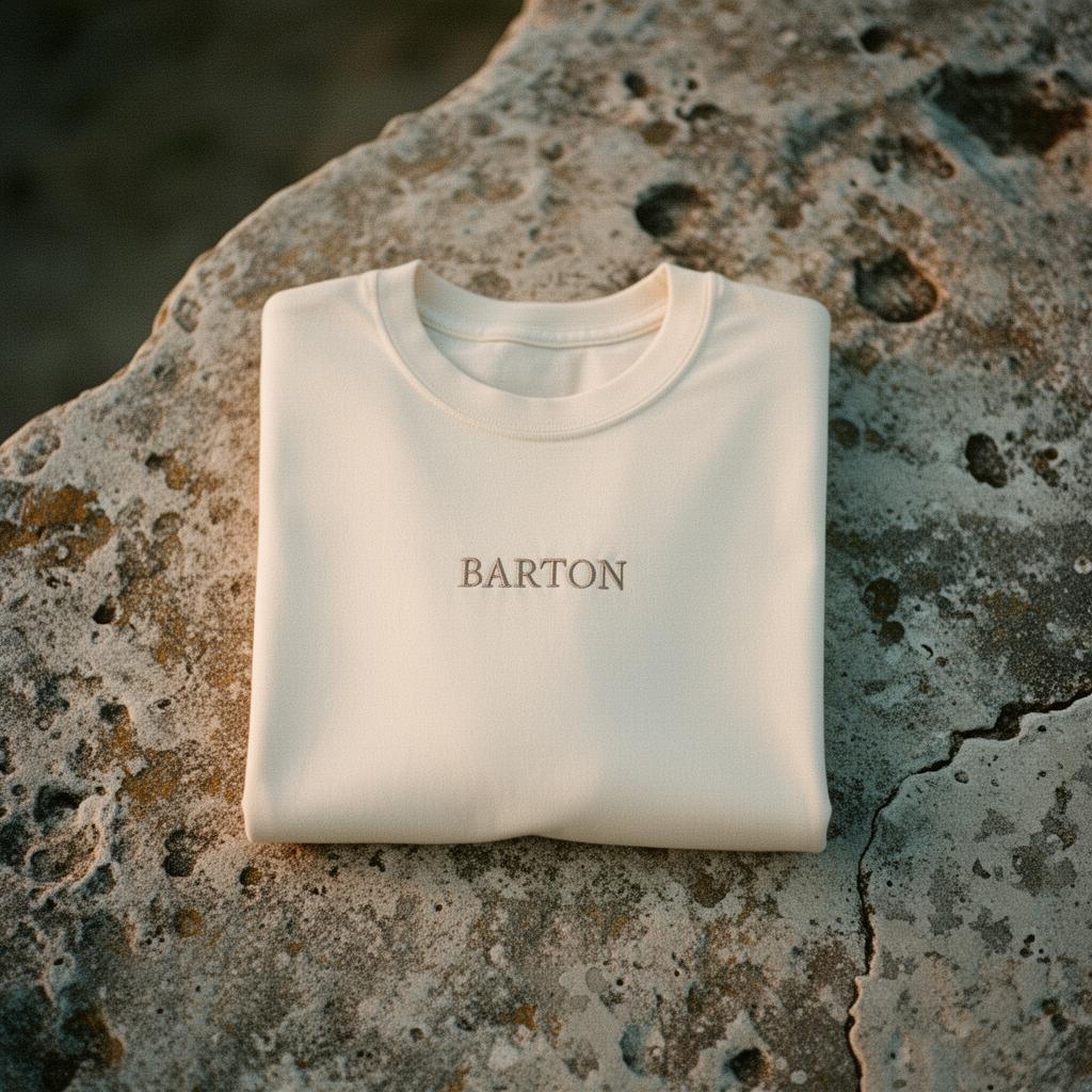 A folded bone heavyweight tee with a small tonal BARTON chest embroidery on weathered limestone.